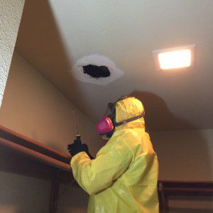 A person in yellow suit and mask spraying water on ceiling.
